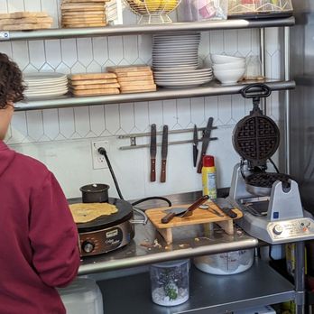 Dek Doi Cafe - A person wearing a red hoodie is cooking a pancake on a black electric griddle in a commercial kitchen with shelves of plates and utensils. - Cafes Near Me - Oakland, California