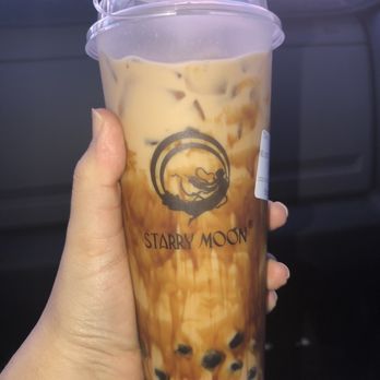 Starry Moon - The Dirty Tea, 100% Sugar, less ice, with boba - A person holds a tall, clear plastic cup of bubble tea from Starry Moon, filled with milk tea and black tapioca pearls at the bottom. The cup features the Starry Moon logo with a silhouette of a girl on a swing under a crescent moon. The background shows the interior of a vehicle, with a blurred view outside the window. - Bubble Tea Near Me - Albuquerque, New Mexico