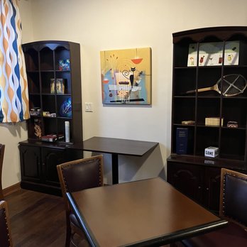North Shore Distillery - Atmosphere and cozy seating! - The image shows a corner of a room with dark wood chairs, a table, and two dark wood bookshelves. A colorful abstract painting hangs on the wall, and patterned curtains are partially visible. - Distilleries Near Me - Green Oaks, Illinois