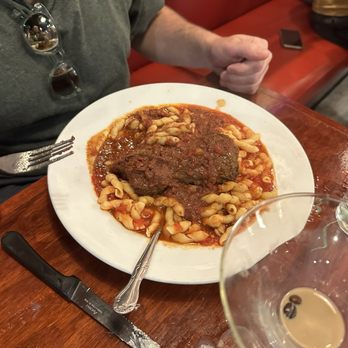 Vinny's Ristorante - Beef Bracciola - A white plate contains cooked pasta topped with a large meatball and tomato sauce. The plate is placed on a wooden table, with a fork and a butter knife beside it. A glass of wine with a few coffee beans at the bottom is partially visible in the foreground. A person wearing a gray shirt and sunglasses hanging from the collar is seated at the table, with a red booth and a smartphone in the background. - Italian Near Me - Somerville, Massachusetts