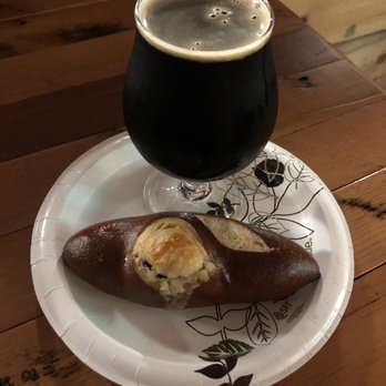High Nine Brewing - Joe stout & Pretzel from 8 Mile Meadow Bakery. - A tulip-shaped glass filled with dark stout beer, featuring a foamy tan head, is positioned on a paper plate. The plate also contains a pretzel with a cheese topping. The setting appears to be a rustic indoor environment with a wooden table surface. - Breweries Near Me - Deep River, Connecticut