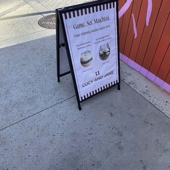 Lucy and Jane - A freestanding sidewalk signboard positioned on a concrete sidewalk advertises a game and drink menu for Lucy and Jane, a business in Boston. The sign features images of a matcha latte and a cocktail, with text promoting their offerings. The background includes a painted wall with purple and pink accents, and the sidewalk shows typical urban elements like a manhole cover and shadows from nearby objects. - Coffee & Tea Near Me - Boston, Massachusetts