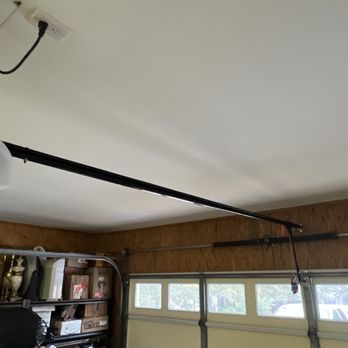 Fowler Overhead Doors - New motor/track. Also Mr. Forest moved the spring on the wall to a stud as it was coming away from the wall. - The image depicts the interior of a garage with a new motor and track system installed for the garage door. The garage door is partially visible, with a wall-mounted spring being moved or adjusted. The garage has a shelving unit with various items, and a garage door opener mechanism is mounted on the ceiling. The wall features a series of small windows allowing natural light into the space. - Garage Door Services Near Me - Benton, Arkansas