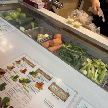 Tbaar - A variety of fresh vegetables including carrots, celery, and leafy greens are arranged behind a glass display at a juice bar or health food store. A menu with colorful images and descriptions is on the counter, and a person is preparing ingredients behind the counter. The setting appears to be a retail or food service environment focused on fresh produce and healthy beverages. - Bubble Tea Near Me - Denver, Colorado