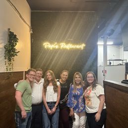 Maria and her family members who were running their restaurant! - Six people stand together inside Papi's Restaurant, a Latin American cuisine establishment. They are positioned in front of a wall with a neon sign that says 'Papi's Restaurant.' The interior features wooden paneling on the lower walls and a dark upper wall with a small plant hanging on the left. The group appears to be enjoying their time, with some smiling and one person holding a phone. The restaurant's kitchen area is partially visible on the right side of the image.