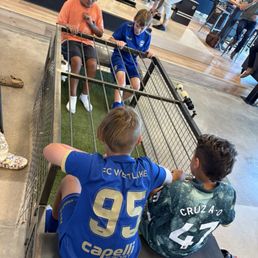 Footy ball (soccer) - Four children, two wearing blue sports jerseys and two in casual clothes, are seated in a small cage playing a game at Pins Mechanical in Austin, TX.
