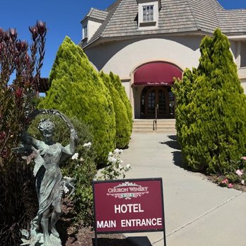 Churon Winery - Front entrance - The photograph depicts the main entrance of Churon Winery hotel, featuring a curved pathway leading to a building with a steep, shingled roof and a maroon awning over the doorway. Flanking the pathway are well-maintained, cone-shaped green bushes. A decorative statue of a girl with a dog is positioned on the left side of the pathway, adding ornamental detail. A sign in front of the statue indicates the hotel main entrance. The sky is clear and blue, suggesting a sunny day. - Wineries Near Me - Temecula, California