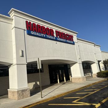 Harbor Freight Tools - The image shows the exterior of a Harbor Freight Tools store with a large sign reading 'Harbor Freight' in red letters and a blue banner underneath that says 'Quality Tools, Lowest Prices.' The building has a white facade with architectural details and a covered entrance. The parking lot in front has marked parking spaces and a curb with a yellow line. The sky is clear and blue. - Hardware Stores Near Me - Margate, Florida