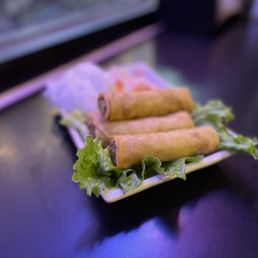 Egg Rolls - A rectangular white plate holds four Vietnamese egg rolls, arranged in a row and garnished with fresh green lettuce leaves. The plate is placed on a dark, smooth table surface, with a blurred background indicating a casual restaurant environment. The image emphasizes the food presentation and the appetizer's appearance.