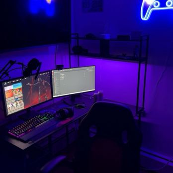 KC Technologies - Dual monitors that KC Tech set up - A computer workspace features two monitors on a desk, with a microphone mounted on a boom arm, gaming keyboard, and headphones. The room is lit with blue and purple LED lighting, creating a gaming or streaming atmosphere. A neon sign of a game controller is mounted on the wall, and there are decorative items and signs on the wall above the desk. An office chair is positioned in front of the desk, and a shelving unit is visible in the background. - IT Services & Computer Repair Near Me - Derry, New Hampshire