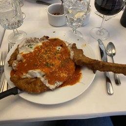 Veal Chop Sinatra - A plate of veal Parmesan with tomato sauce, melted cheese, and herbs, accompanied by a breaded cutlet, on a white table with drinks and utensils.