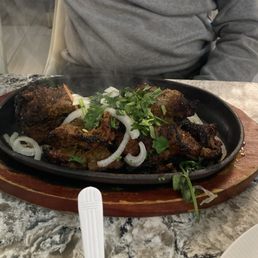 Kebab - A hot cast iron skillet with grilled meat, topped with sliced onions and cilantro, placed on a wooden serving board on a marble table.