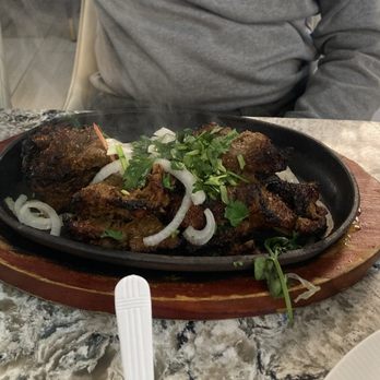 Dera - Kebab - A hot cast iron skillet with grilled meat, topped with sliced onions and cilantro, placed on a wooden serving board on a marble table. - Pakistani Near Me - Springfield, Virginia