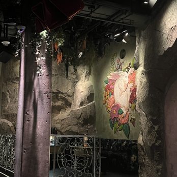 Caveau - Atmosphere - The image shows an indoor space with rock-textured walls and a small stage area. A mural of a rabbit surrounded by colorful flowers decorates one wall. Overhead, there are stage lights and some hanging greenery. The space appears to be a restaurant or bar with a whimsical, natural theme, including a decorative metal railing and a few chairs stacked upside down near the ceiling. - Lounges Near Me - Boston, Massachusetts