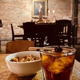 a table with a bowl of food and a drink - A glass filled with iced beverage, topped with a cherry, is positioned on a rustic wooden table. Next to it is a white bowl containing a snack mix with nuts and possibly dried fruit. The background features a brick wall, a framed portrait, and a dining area with dark wooden chairs and a table, suggesting a cozy, vintage-style restaurant or cafe environment.