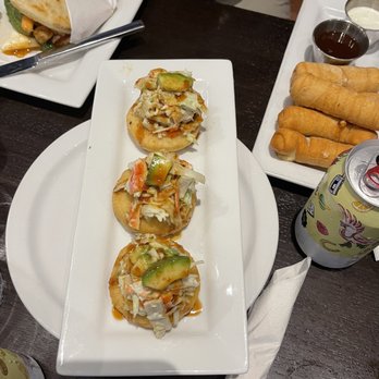 Bienmesabe - Three Venezuelan arepas topped with chopped vegetables, such as cucumbers, tomatoes, and cabbage, are arranged on a rectangular white plate. The arepas are garnished with a sauce, likely a dressing or condiment. The plate is placed on a dark wooden table, with other dishes, drinks, and condiments visible nearby, including spring rolls, dipping sauces, and canned beverages. - Venezuelan Near Me - Chicago, Illinois