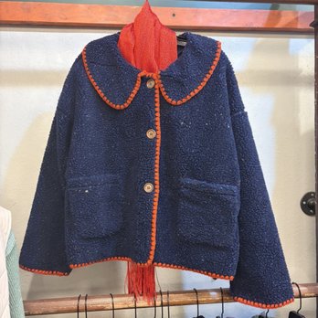 Camp Willow - Wish they had it in our size!! So cute. - A navy blue fleece jacket with orange trim along the collar, front opening, and hem is hanging on a clothing rack. The jacket features two front pockets and four buttons. A red scarf is draped underneath the jacket, visible at the collar. The background includes a wall with a colorful sign and other clothing items hanging below the jacket. - Women's Clothing Near Me - Park Ridge, Illinois