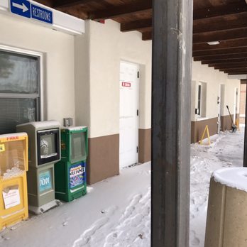 La Bajada Rest Area - Snow covers the sidewalk outside a building with newspaper boxes, a restroom sign, and a door, with snow shovels visible in the background. - Rest Stops Near Me - Santa Fe, New Mexico