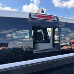 A woman wearing sunglasses leans on her arms inside a vehicle, with a reflection of the sky and clouds visible on the window.