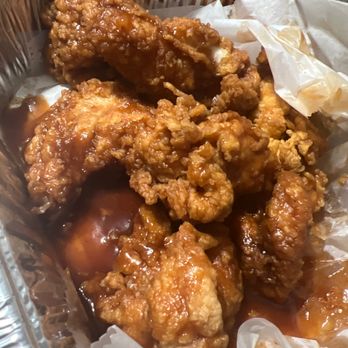 Wings Over Worcester - The image displays several pieces of fried chicken wings coated in a glossy sauce, placed in a disposable foil container lined with paper. The wings appear crispy and are covered with a sauce that has a shiny, sticky appearance. The container is partially open, revealing the food inside, and is suitable for takeout or delivery. - Chicken Wings Near Me - Worcester, Massachusetts