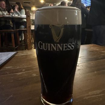 Murphys Irish Pub - Guinness :) - A tall glass of Guinness stout with a creamy head is placed on a wooden table inside a pub, with patrons and a menu visible in the background. - Irish Pub Near Me - Murphys, California