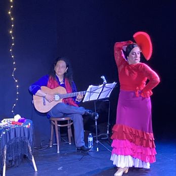 Teatro Ecas - Flamenco - A flamenco dancer dressed in a red top and a long purple skirt with ruffles performs on stage, striking a pose with one arm raised and the other on her hip. A guitarist, seated on a stool, plays an acoustic guitar nearby. The stage background is dark, with a string of small lights visible on the left side of the image. A small table with colorful decorations is also present on the stage. - Performing Arts Near Me - Providence, Rhode Island