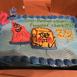 Custom 1/4 Sheet Cake - The cake has a blue icing base with piped border decoration around the edges. It features cartoon characters resembling SpongeBob SquarePants and Patrick, made with colorful icing. There are candles shaped as the numbers 2 and 5, indicating a 25th birthday celebration. The message written in green icing says, "You Know What's Funnier Than 2? 25!" The cake appears to be in a cardboard box, ready for celebration.