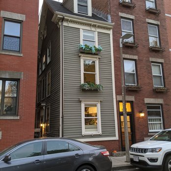 Skinny House - A narrow, three-story house with gray horizontal siding and white trim, situated between two larger brick buildings. The house has three visible windows, each with flower boxes containing greenery and flowers. The top floor features a small dormer window with a gabled roof. The entrance is on the side, with a small pathway leading to the door. Cars are parked in front on the street, and a streetlamp is nearby. The setting appears to be in a city neighborhood with historic architecture. - Landmarks & Historical Buildings Near Me - Boston, Massachusetts