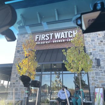 First Watch - Outside - The image shows the front of a First Watch restaurant with two people sitting on a bench outside, surrounded by trees. - Breakfast & Brunch Near Me - Newark, Delaware