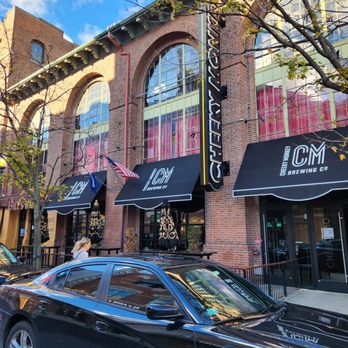 Cheeky Monkey Brewing Company - The image shows the front of Green Monkey Brewing Co., a brewpub with brick architecture, large arched windows, and black awnings displaying its logo. A black car is parked in front, and a person is walking nearby. - Venues & Event Spaces Near Me - Boston, Massachusetts