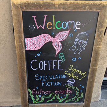 Beastly Books - A chalkboard sign with a wooden frame displays colorful handwritten text and illustrations. It reads 'Welcome' in multicolored letters, with drawings of a pink mermaid tail, jellyfish, and seaweed, indicating a theme related to the ocean. The sign advertises coffee, signed copies, speculative fiction, and author events, suggesting it is for a bookstore or literary venue that hosts community activities and sells books. The sign is placed outdoors against a beige wall. - Bookstores Near Me - Santa Fe, New Mexico