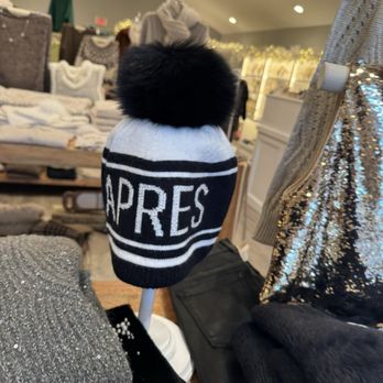 Valentina - A knit hat with a large black pom-pom on top, predominantly white with black accents, displaying the word 'APRES' in bold, capital letters. The hat is placed on a white mannequin head in a retail store setting, surrounded by clothing and accessories, with a chandelier and store fixtures visible in the background. - Women's Clothing Near Me - Glencoe, Illinois