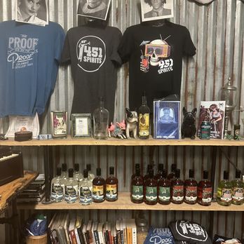 451 Spirits - The shop has a punk rock feel. - The image shows a display shelf set against a corrugated metal wall, featuring three T-shirts with printed graphics and photos of people's faces, bottles of spirits, small figurines, books, and other collectibles. The top row includes T-shirts with logos and artwork, each with a photo of a person's face as a mask. The middle shelf holds bottles of spirits, small figurines, and framed items. The bottom shelf contains books, folded clothing, and additional items. The setting appears to be a retail or display area for a distillery or related business. - Distilleries Near Me - Columbus, Ohio