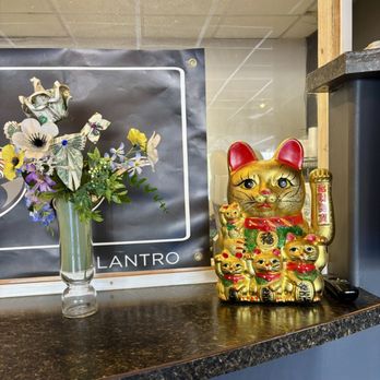 Bamboo by Kulantro - deocrations - A gold-colored Maneki-neko figurine with multiple smaller cats is positioned on a dark, speckled countertop. The figurine features a large cat with a raised paw, and several smaller cats around it, some holding objects like a coin or a scroll. To the left, there is a glass vase containing a mixed flower arrangement with various flowers and greenery. The background includes a black sign with white text and a partial view of a wall and ceiling. - Vietnamese Near Me - Albuquerque, New Mexico