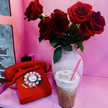 Jasmine Jo - The image features a pink table with a white ribbed vase containing red roses, a vintage red rotary phone, and a clear glass of iced coffee with ice cubes and a pink straw. The background includes a pink wall and a window with a brick wall visible outside. The scene combines floral decor with retro and beverage elements, creating a colorful and eclectic setting. - Coffee & Tea Near Me - Tempe, Arizona