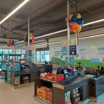 ALDI - Cashiers - The image shows the interior of an ALDI grocery store in Washington, DC, with checkout counters numbered 3, 4, and 5. Balloons are attached to poles above the counters, and large wall graphics promote sustainability initiatives like reducing paper and plastic use. The store has bright lighting, large windows, and various products near the checkout area. Two staff members are visible, one attending to the register and another organizing items in a shopping cart. - Grocery Near Me - Washington, DC, District of Columbia