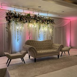 A stage setup featuring a floral arch, draped curtains, and vintage-style beige sofa and chairs, suitable for weddings or special events.