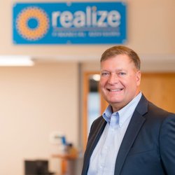 Realize Financial Advisors - A headshot of a team member in a dark blazer standing in the entrance area of Realize Financial Advisors with the bright blue...