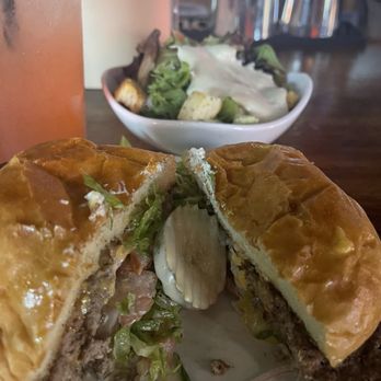 Sunset Lounge - Sunset Smash Burger with Side Salad - A burger with a toasted bun, lettuce, pickles, and sauce, accompanied by a side salad with dressing and a pink beverage in a glass. - Burgers Near Me - Norwalk, Iowa