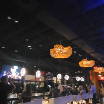 Morse Code Lounge - A dimly lit bar with hanging wooden light fixtures, patrons seated at the bar, and a dark ceiling with blue lighting accents. - Wine Bars Near Me - Winter Park, Florida