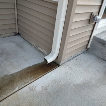 Shodeen - The image depicts the exterior corner of a house with beige vinyl siding, a white plastic downspout attached to the wall, concrete steps leading to a door, and a concrete porch area. The ground shows some discoloration and staining, and the siding meets the concrete foundation at the bottom. The scene is well-lit, indicating daytime. - Real Estate Services Near Me - Geneva, Illinois
