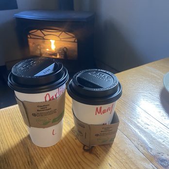 Bistro Espresso - Worm coffee - Two disposable coffee cups with black plastic lids and cardboard sleeves are positioned on a wooden table. One cup is larger, and the other is smaller, both labeled with red handwriting. The background features a fireplace with a visible flame, creating a warm ambiance in the indoor space. - Coffee & Tea Near Me - Arnold, California