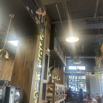 Black Lion Cafe - Pastry bar and ordering line - The image shows the interior of a café with a modern design, including wooden wall panels and hanging pendant lights. There are several customers seated at tables and near the window, some working on laptops. A menu board is visible on the wall, and the space is illuminated by natural light from large windows at the front. The ceiling has exposed ductwork and industrial-style lighting fixtures. - Cafes Near Me - Silver Spring, Maryland