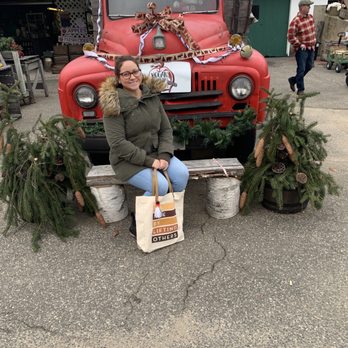 Hogan's Cider Mill - Cute places for photos - A woman wearing glasses, a green coat with a fur-lined hood, and light blue jeans sits on a wooden bench made of logs in front of a vintage red truck decorated for the holiday season. The truck has a large bow on its hood and is adorned with greenery and pinecones. The setting appears to be an outdoor market or festive display area, with other people and market stalls visible in the background. - Venues & Event Spaces Near Me - Burlington, Connecticut