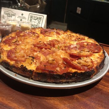 Cape Cod Cafe - Pepperoni and onion with laced edges - A pizza featuring pepperoni and onions is placed on a dark wooden table at Cape Cod Cafe. The pizza has a slightly charred crust and is topped with melted cheese, pepperoni slices, and cooked onions. In the background, there are small containers of dipping sauces and a QR code sign, indicating a casual dining setting. The cafe's interior includes a bar area with bottles and glasses visible. - Pizza Near Me - Brockton, Massachusetts