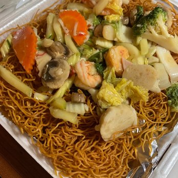 Bamboo by Kulantro - The image shows a styrofoam takeout container filled with crispy fried noodles as the base. Topping the noodles are various vegetables such as broccoli, mushrooms, carrots, and cabbage, along with seafood including shrimp. The dish appears to be a seafood stir-fry served over fried noodles, typical of Asian cuisine. In the background, there are additional food containers and utensils, indicating a takeout or delivery meal. - Vietnamese Near Me - Albuquerque, New Mexico