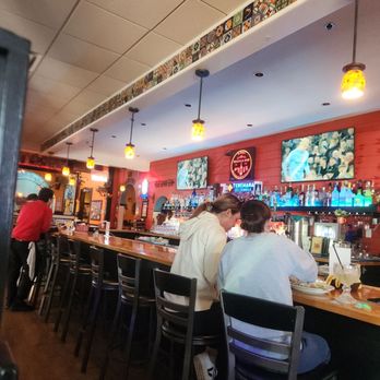 Los Cantaros - Two women sit at a bar counter in a restaurant with TVs showing sports, colorful wall decor, and hanging pendant lights. - Mexican Near Me - Hampton, New Hampshire
