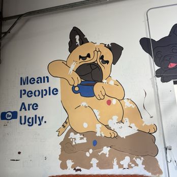 Just Be Coffee Roasters - The image shows a mural of a cartoon dog with a sad expression, wearing a blue collar, sitting on a rock. The mural is painted on a white wall, with some white paint or damage visible on the artwork. To the left of the mural, there is text that reads 'Mean People Are Ugly.' The setting appears to be indoors, possibly in a cafe or creative space, with a brown leather couch at the bottom of the image and some visible piping and structural elements. - Coffee Roasteries Near Me - Munster, Indiana