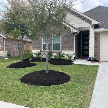 Seventree Landscaping - the front yard of a home - The image shows a residential front yard with two trees, mulched flower beds, and a concrete driveway leading to a garage, in front of a brick house. - Lawn Services Near Me