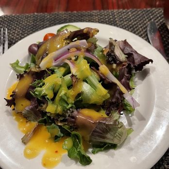 Nuovo - House salad - A fresh mixed green salad featuring various leafy greens, sliced red onions, and a yellow salad dressing drizzled over the top. The salad is presented on a white plate placed on a woven placemat, with a wooden table surface visible in the background. The setting appears to be a restaurant dining environment. - Italian Near Me - Worcester, Massachusetts